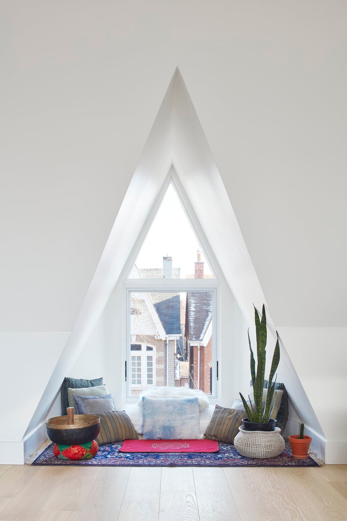 Cozy attic nook with triangular window, cushions, plants, and colourful rug.