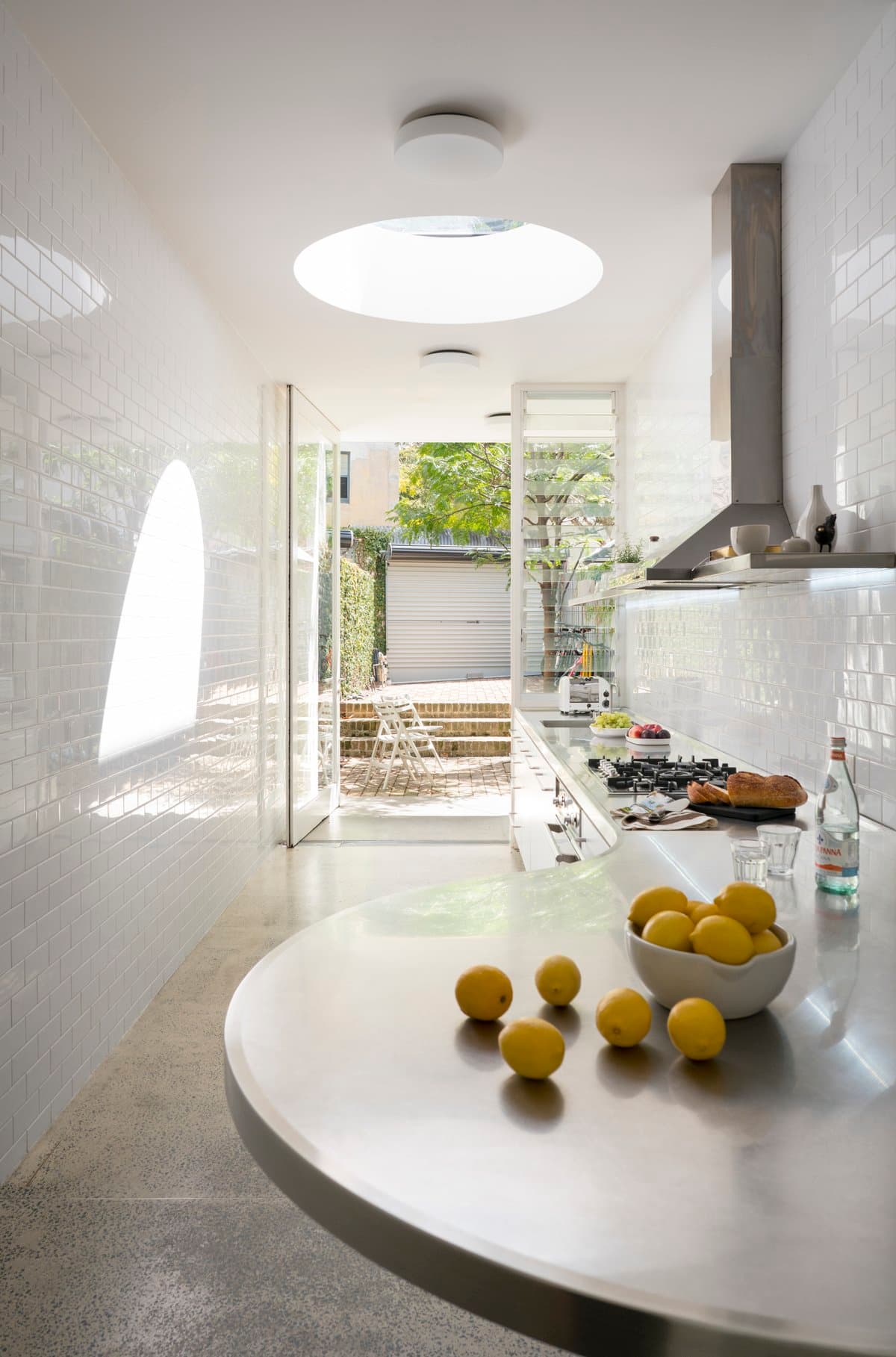 Modern minimalist kitchen with circular skylight, white subway tiles, and floor-to-ceiling glass doors.