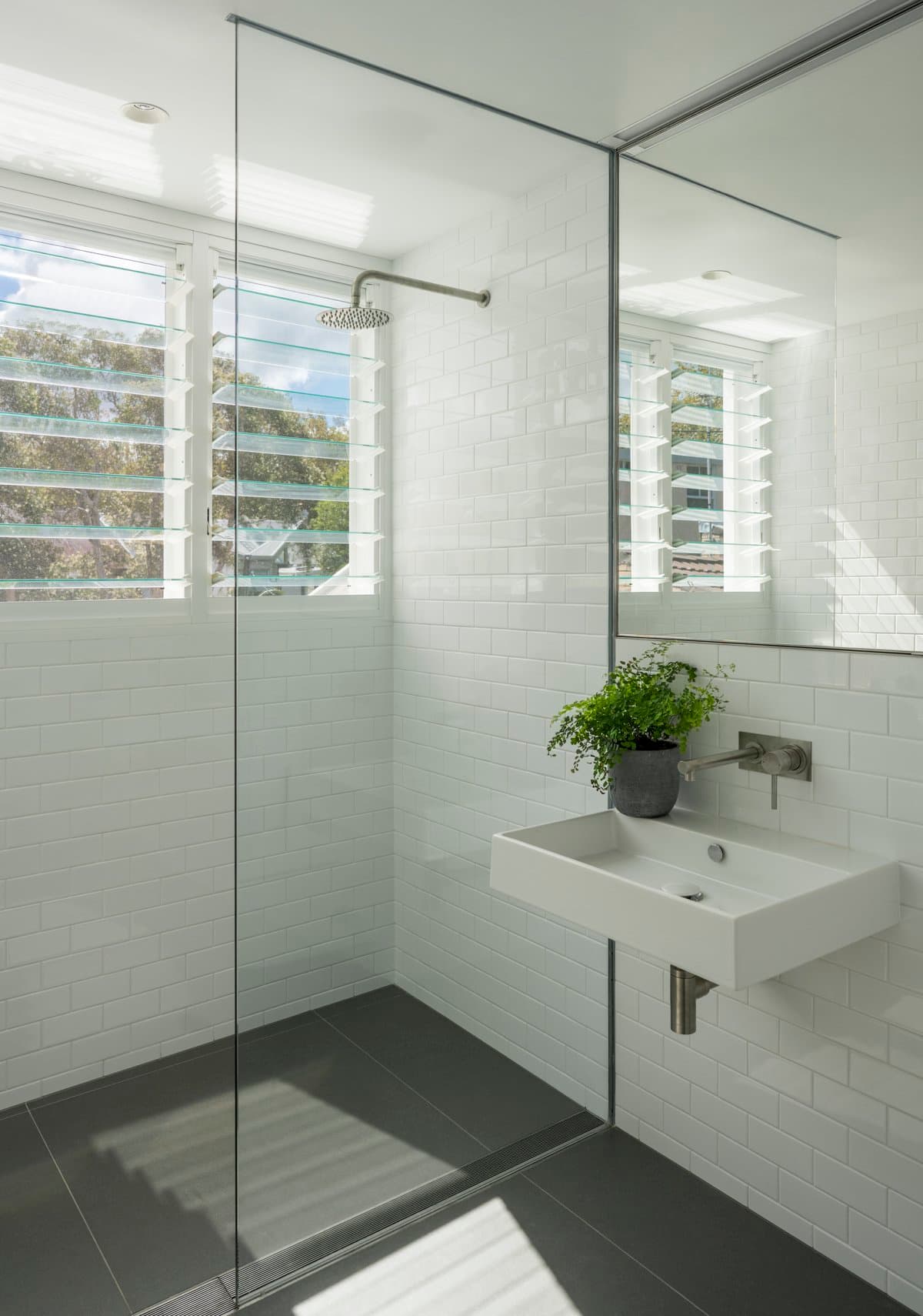 Modern, minimalist bathroom with glass shower, wall-mounted sink, and natural light from white plantation shutters.