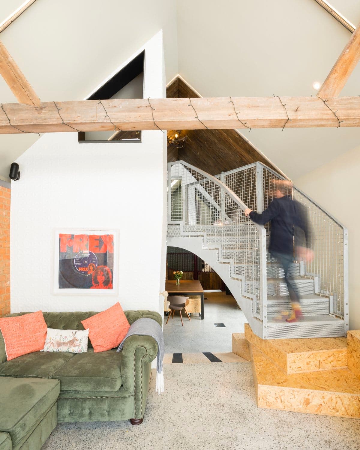 Modern living room with green sectional, industrial staircase, abstract art, and rustic wood beams.