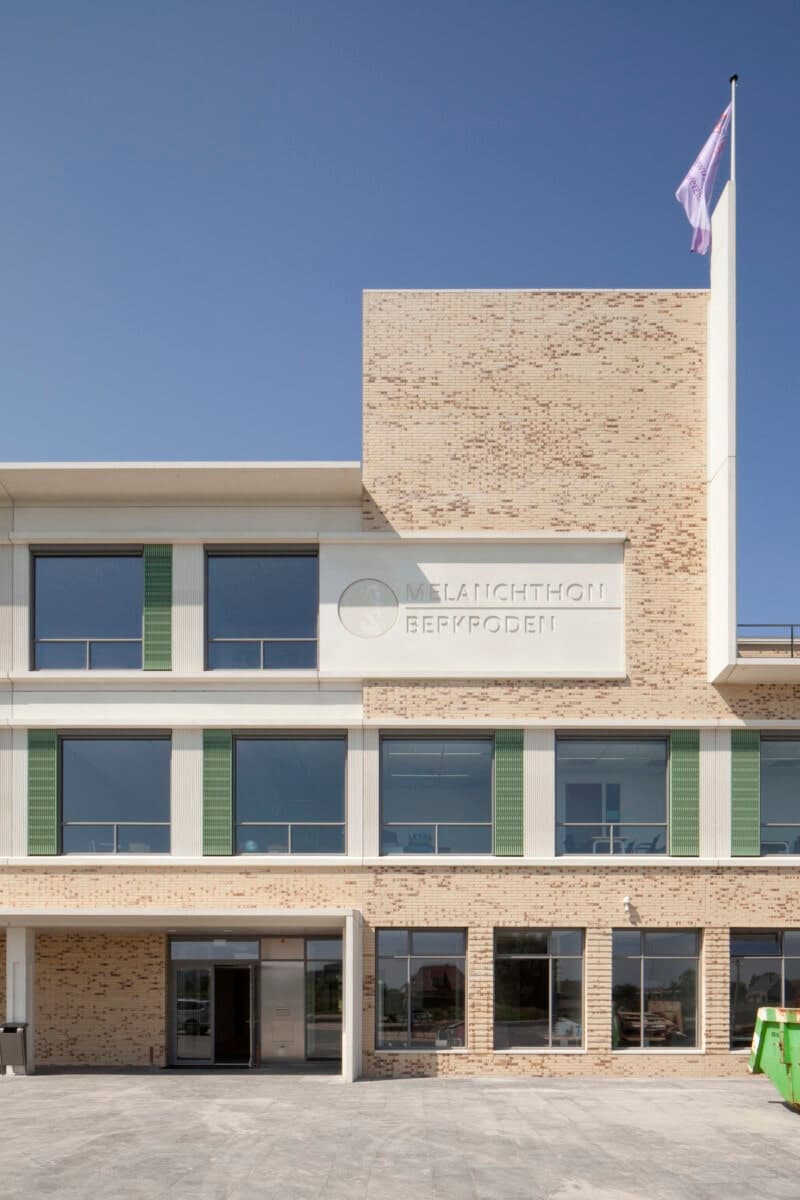 Modern three-story brick building with large windows, flagpole, and green panels: VMBO Melanchton Berkroden.