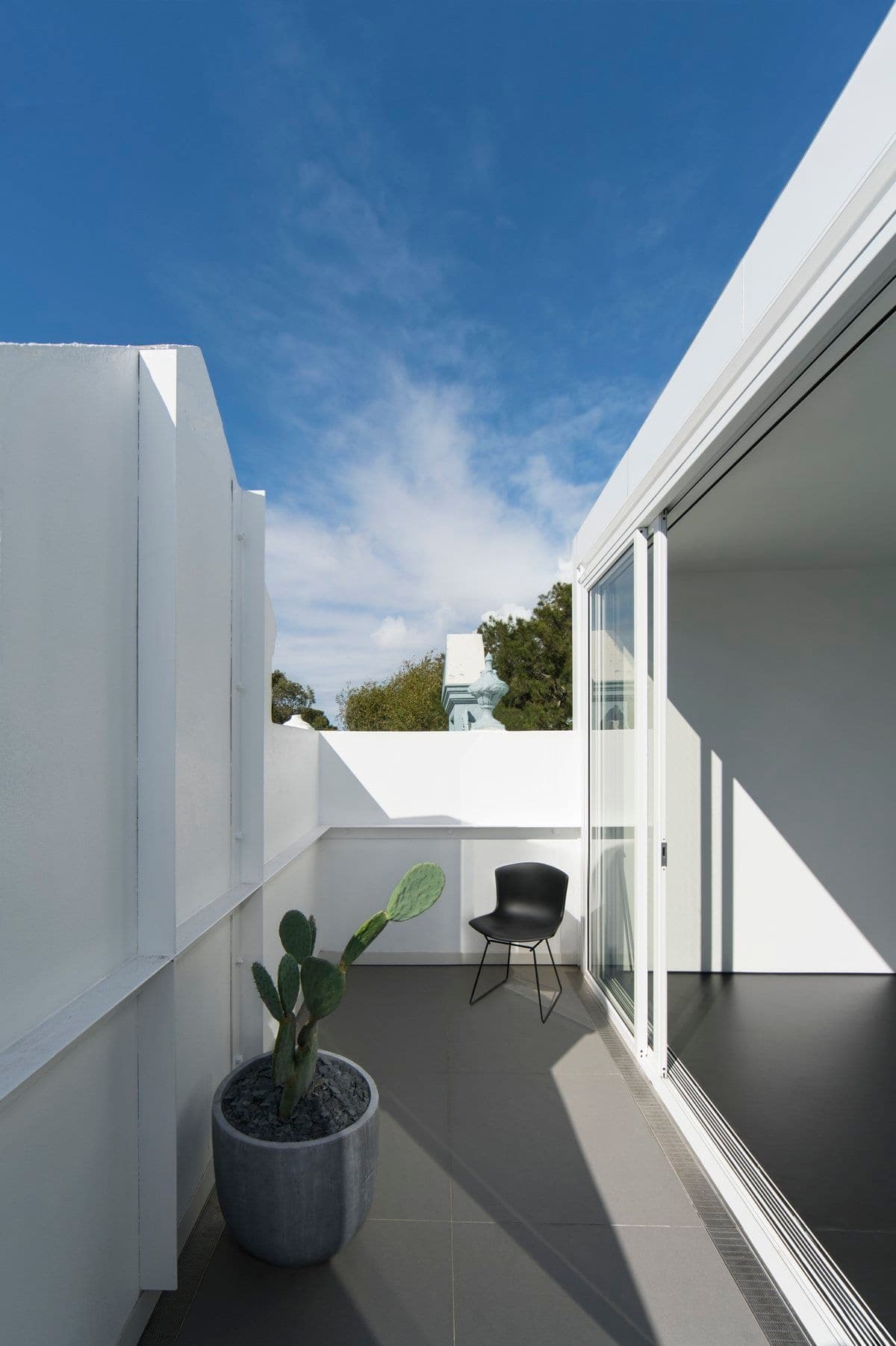 Modern minimalist outdoor terrace with white walls, black chair, cactus, and sliding glass door.
