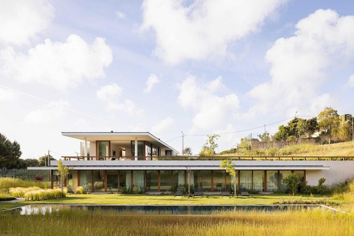 Modern two-storey house in lush landscape with pond, large windows, and hill background.