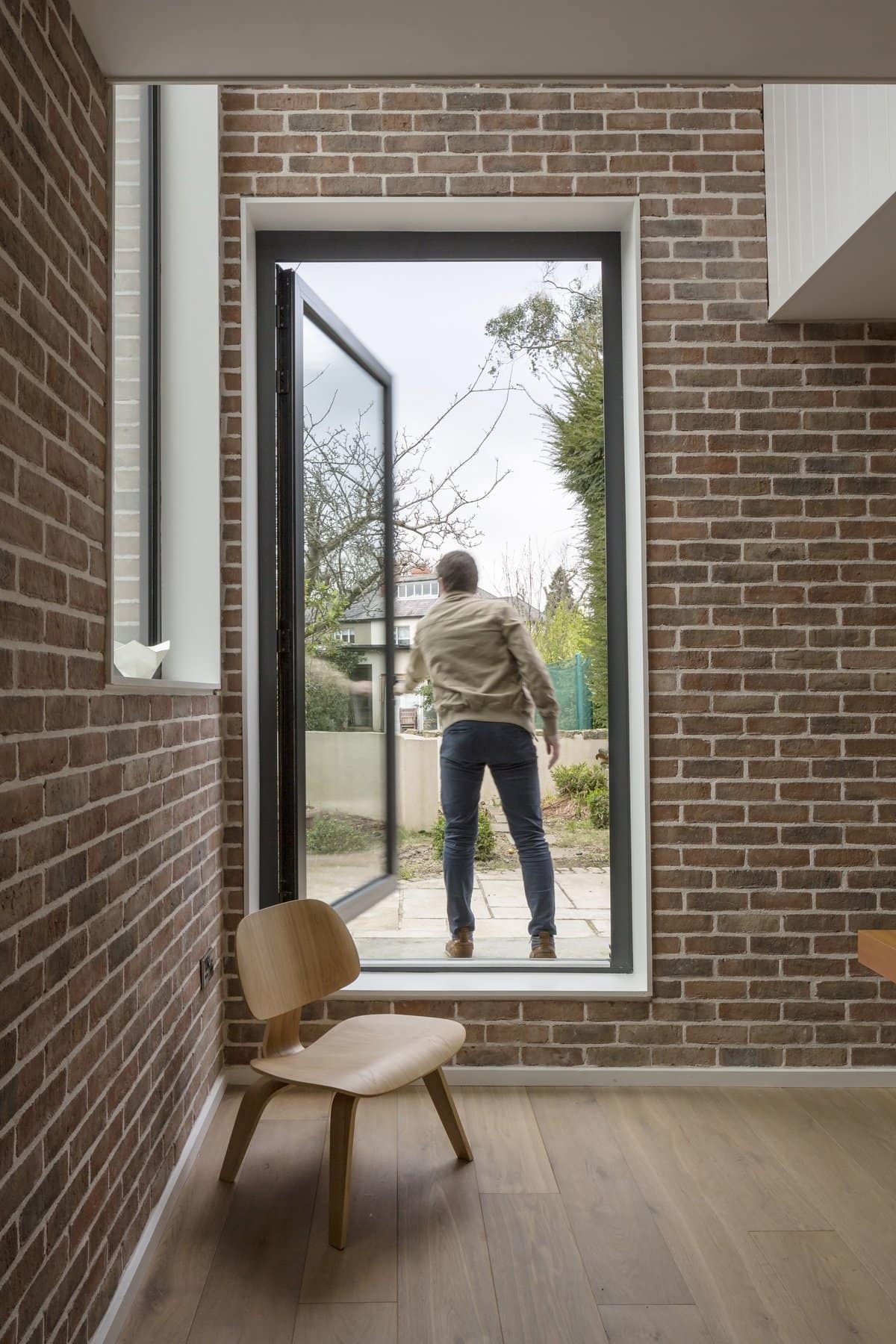 Modern interior with floor-to-ceiling window, minimalist chair, and person in garden outside.