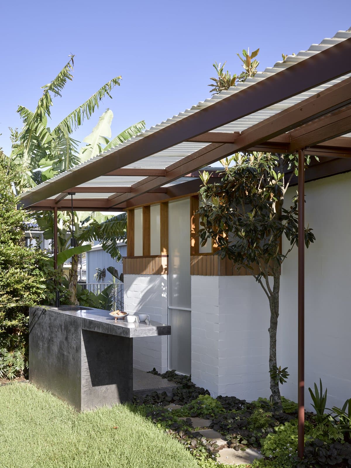 Modern outdoor kitchen with sleek stone countertop, white walls, wooden accents, and lush greenery.