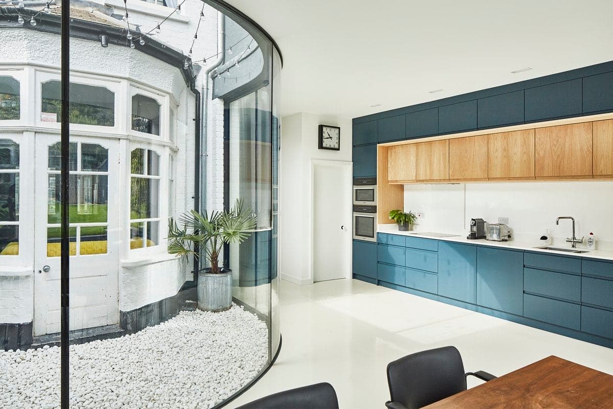 Modern kitchen with curved glass wall and outdoor space, wooden and blue cabinets, natural light.
