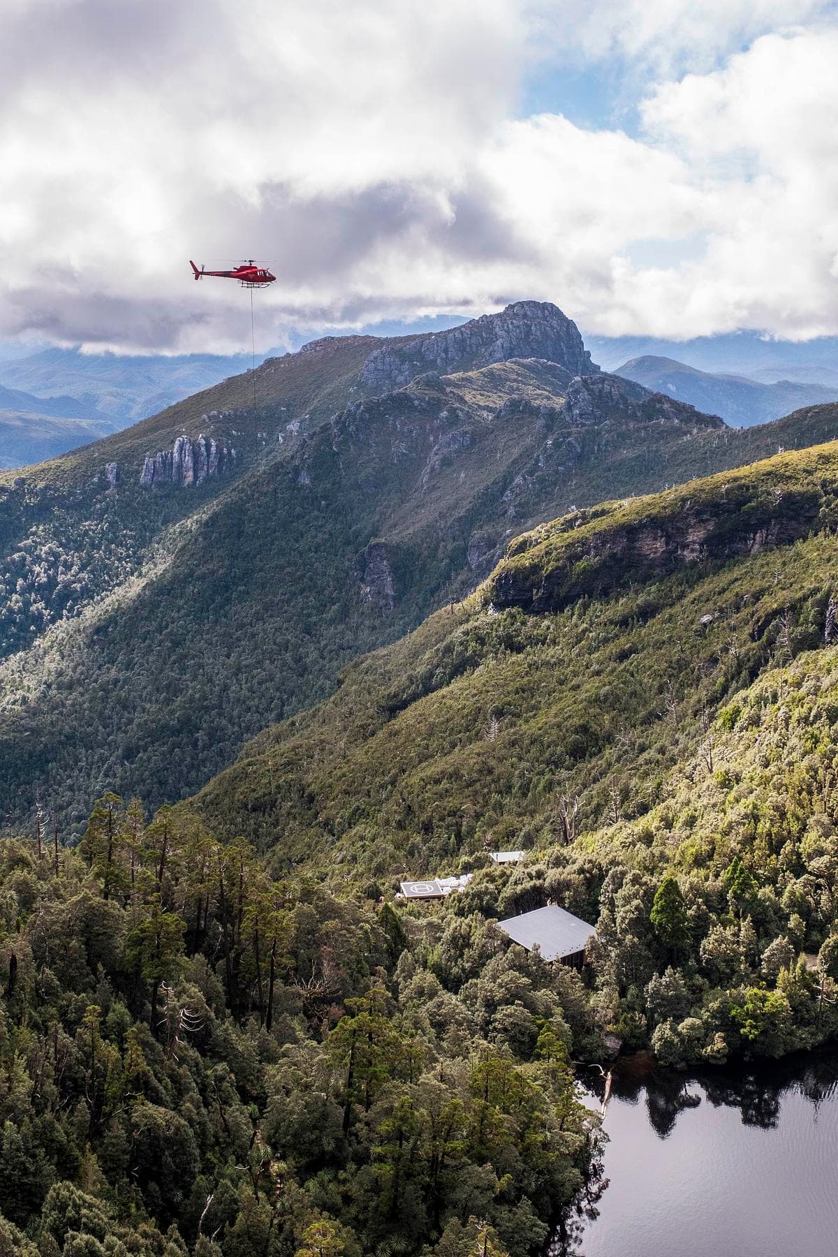 A helicopter above the remote mountain site, with the hut visible beside the lake.