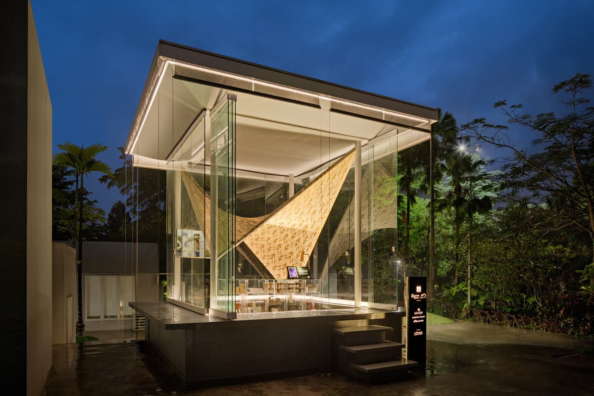 The glazed pavilion and its suspended woven form are illuminated at dusk.