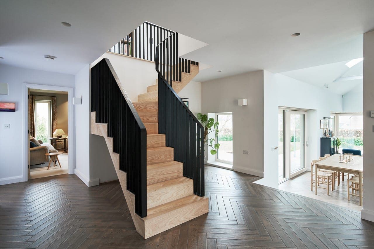 Ash staircase with a black metal balustrade rising from a herringbone-floored hall