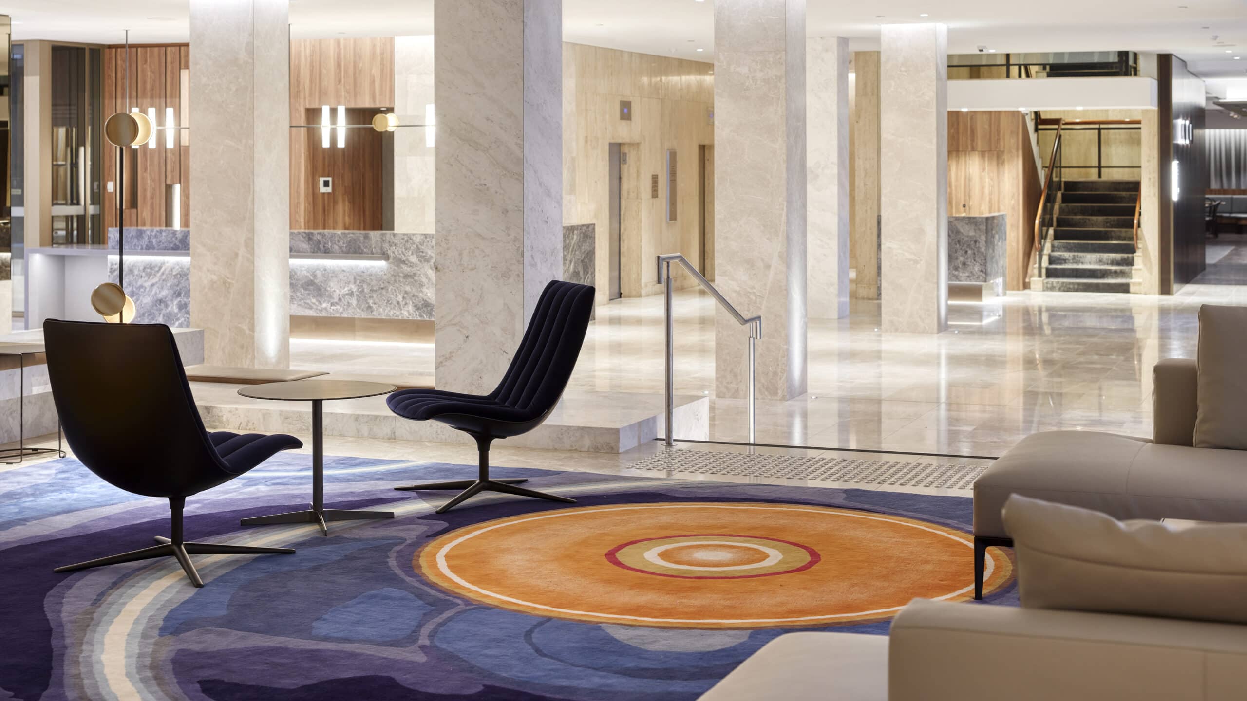 Hotel lobby with pale stone columns, timber wall panels and seating around a colourful circular-patterned rug.