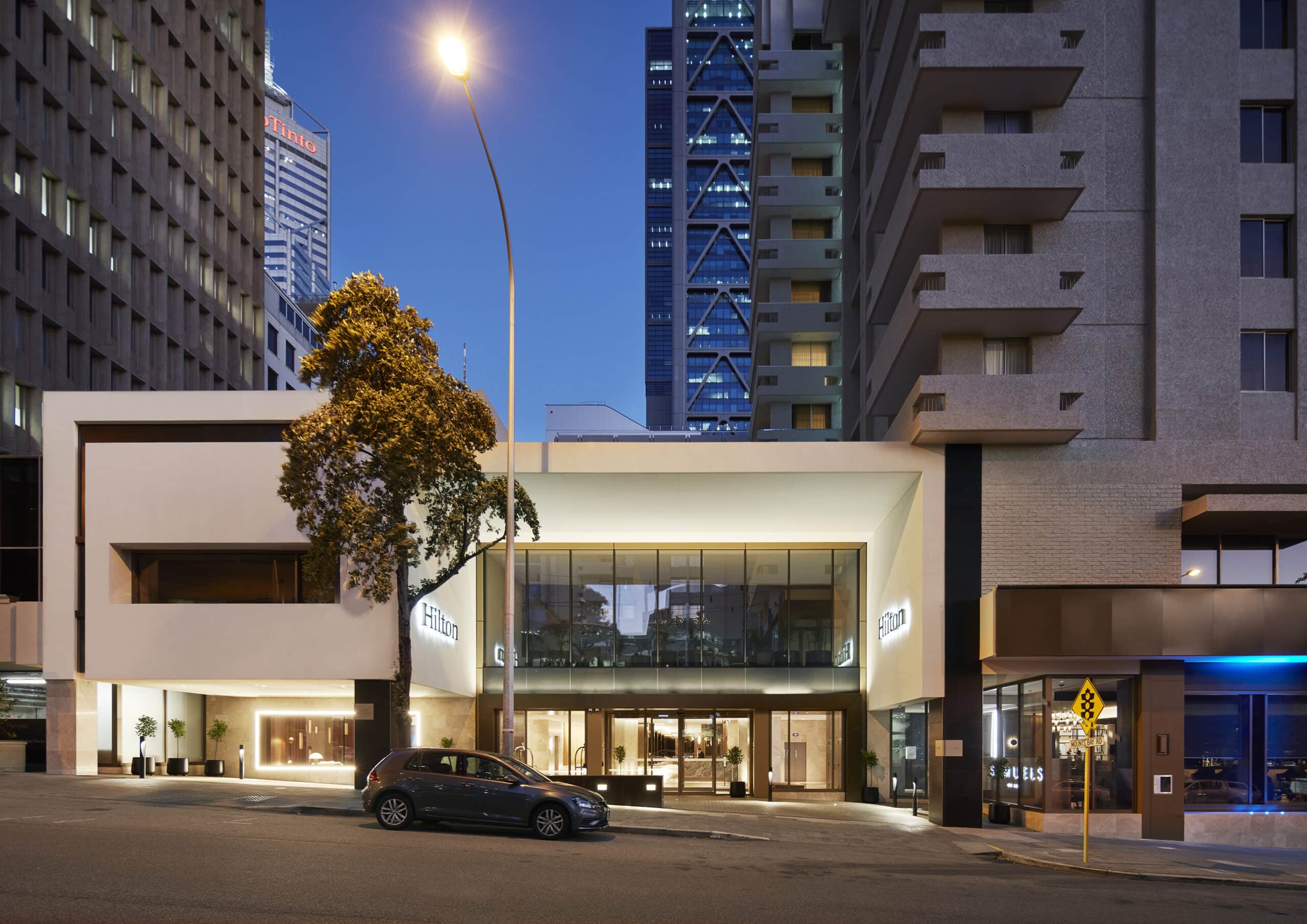 Illuminated hotel entrance and glazed upper-level facade viewed from the street at dusk.