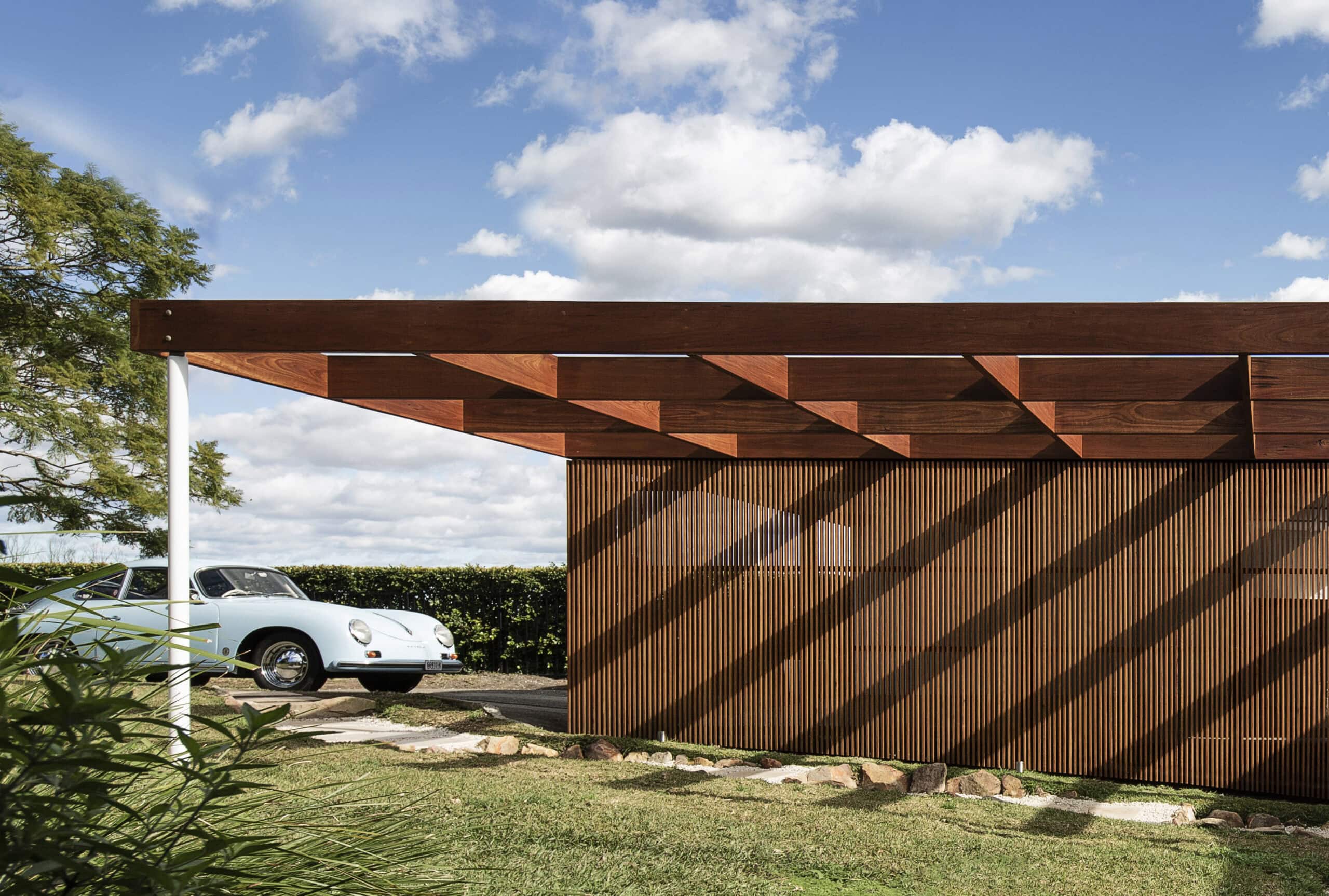 Low timber-clad residence with a projecting exposed-beam canopy beside a landscaped driveway.