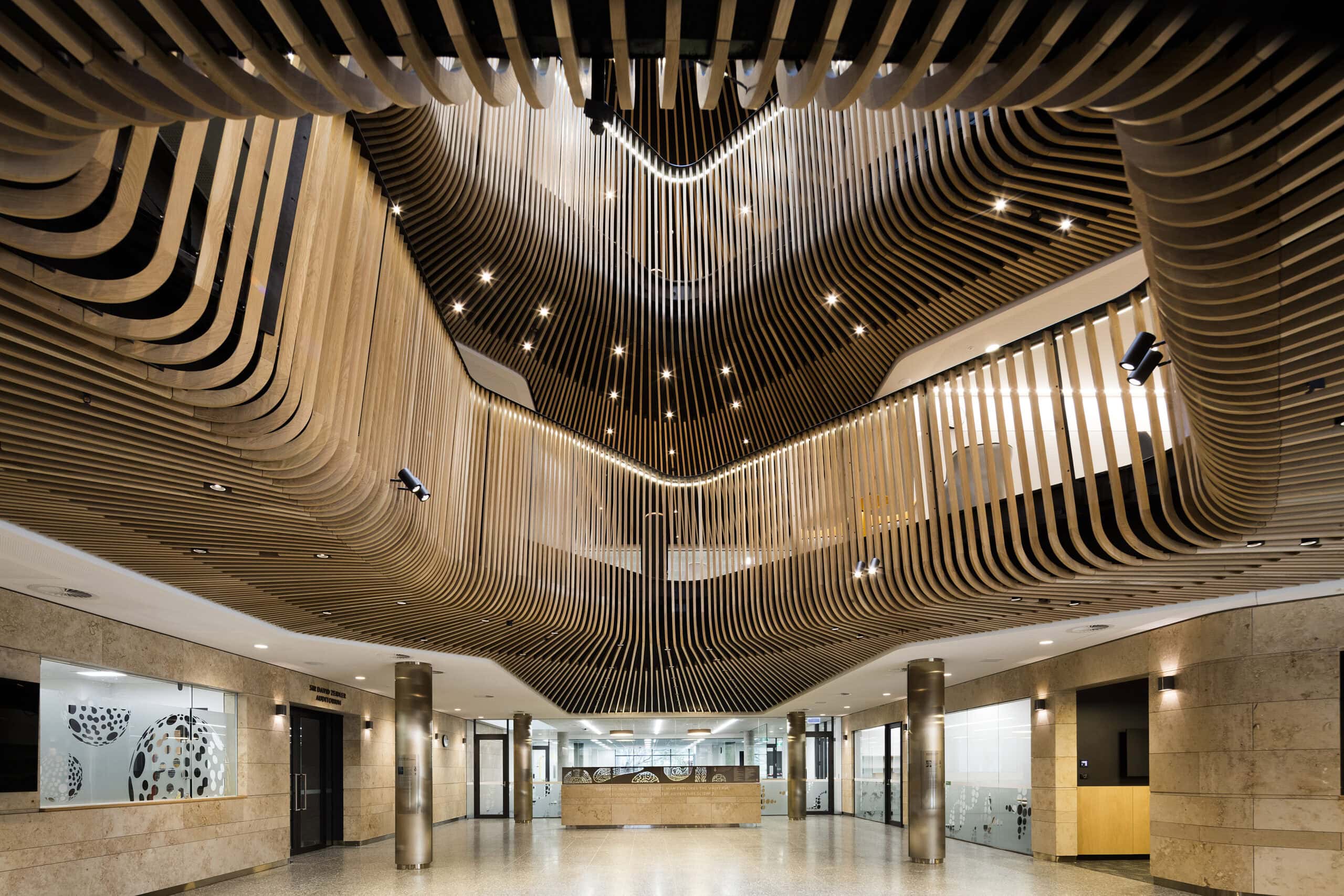 Atrium framed by curved timber slats on multiple levels and suspended vertical light elements.
