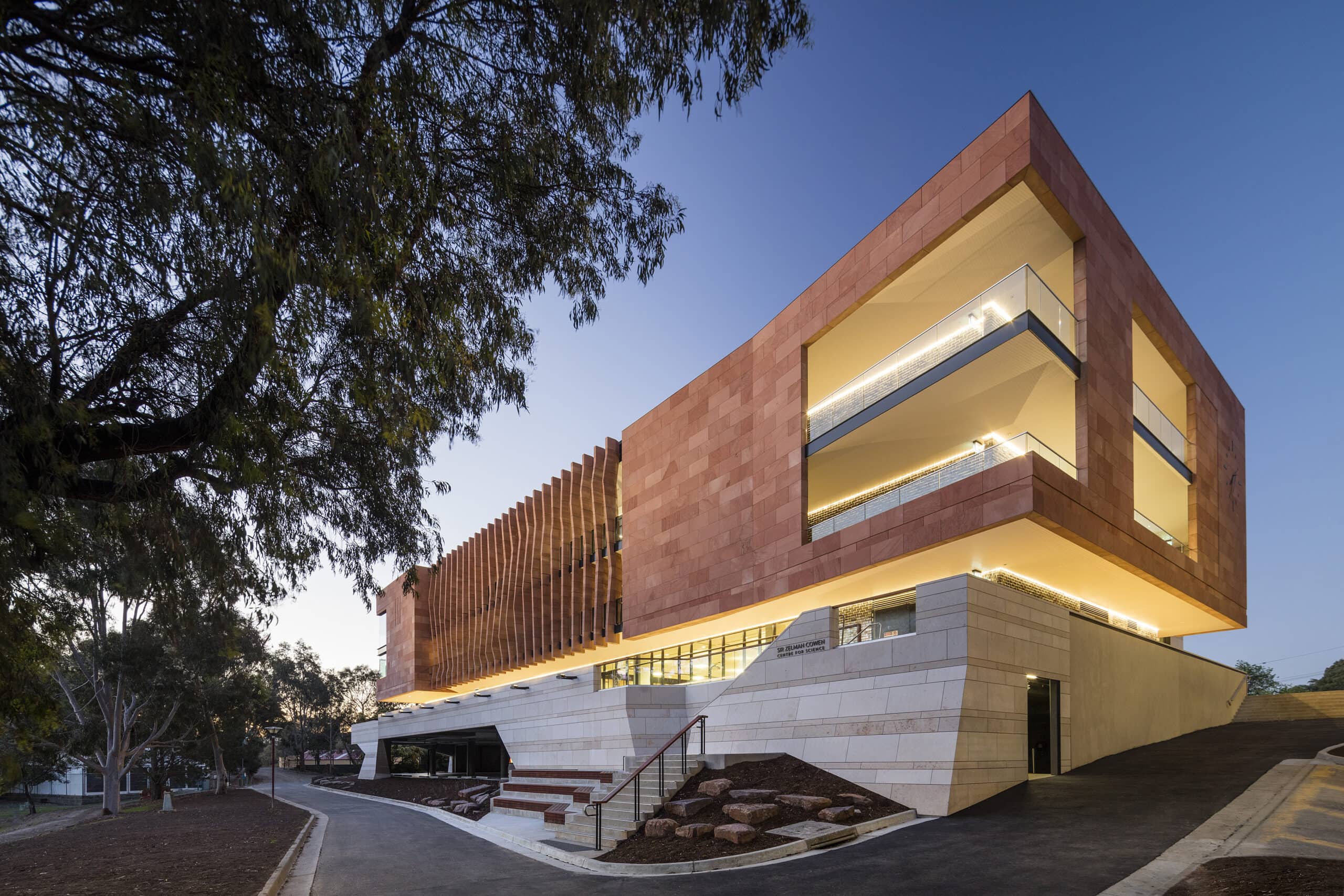 Angular stone-clad science building with illuminated horizontal glazing beside trees at dusk.