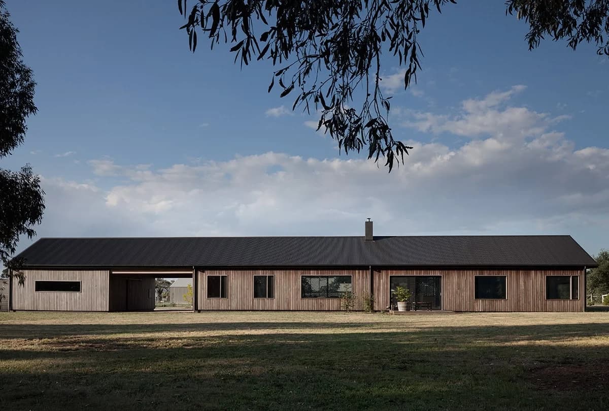 Long single-storey timber house with a dark pitched roof, sheltered openings and a broad lawn.
