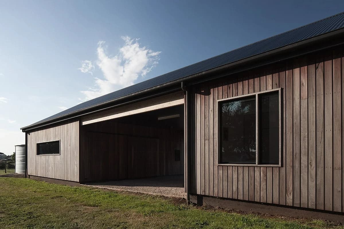 Timber facade with recessed openings and a sheltered central passage beside a lawn.