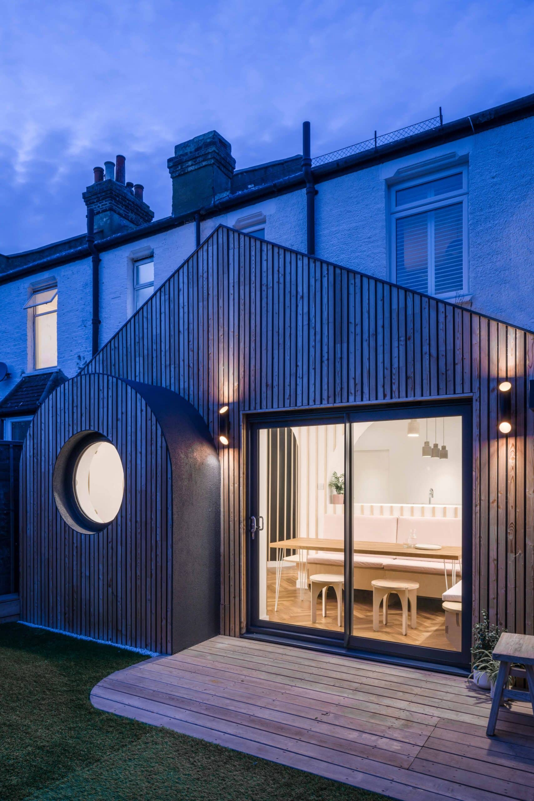 Timber-clad rear extension with a round window, pitched roof and illuminated dining nook at dusk.