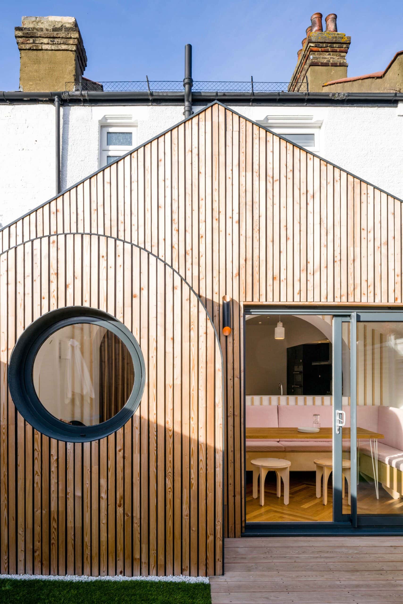 Sunlit timber facade with a circular window and sliding glass doors opening to a dining nook.