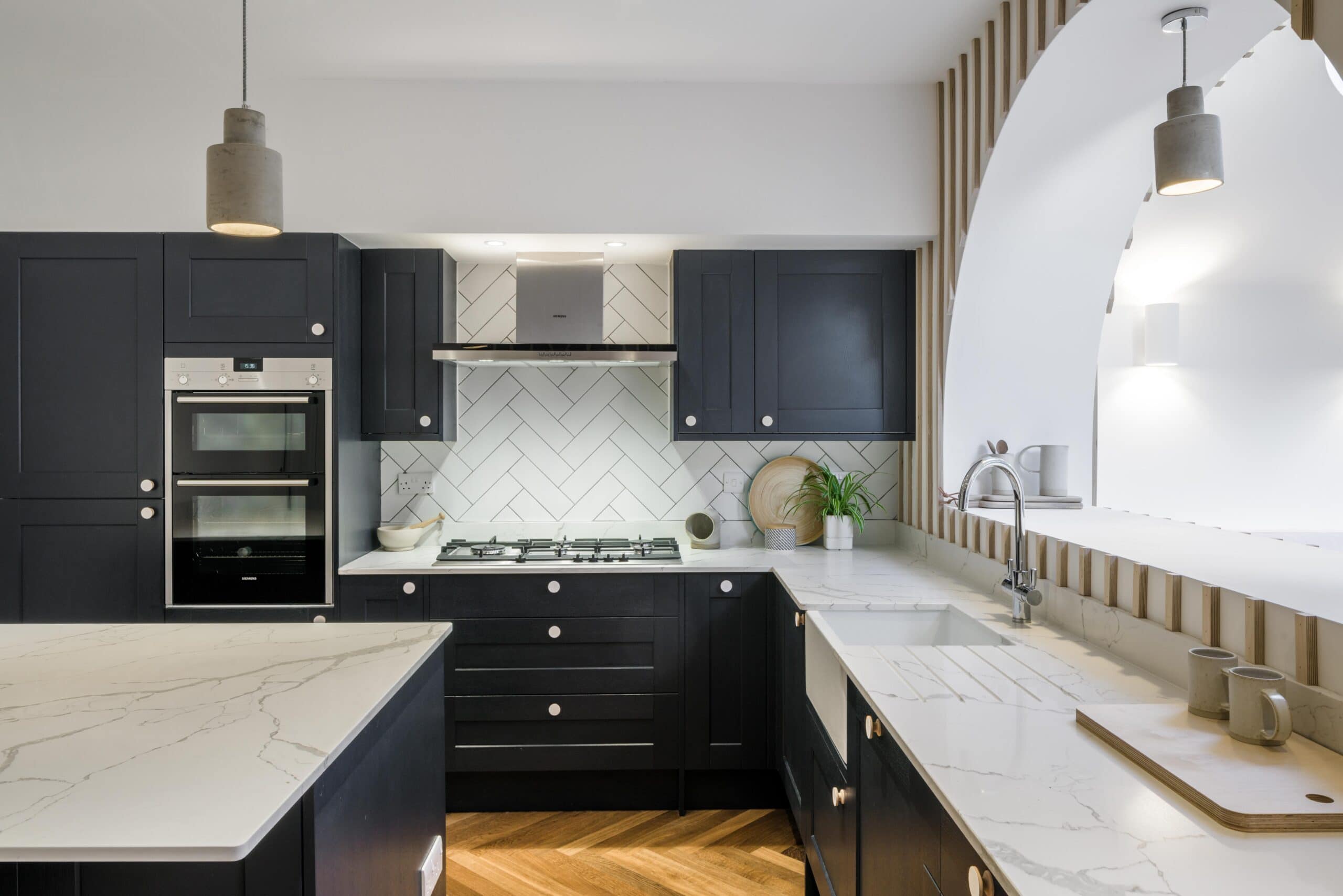 Dark kitchen cabinetry and white worktops beside a herringbone splashback and arched opening.