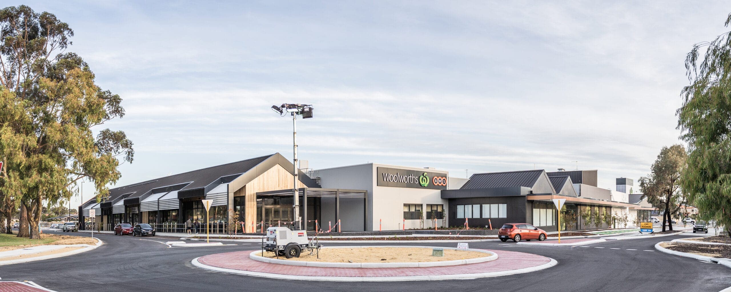 Completed shopping centre frontage with pitched roof forms, entrance glazing and a roundabout in front.