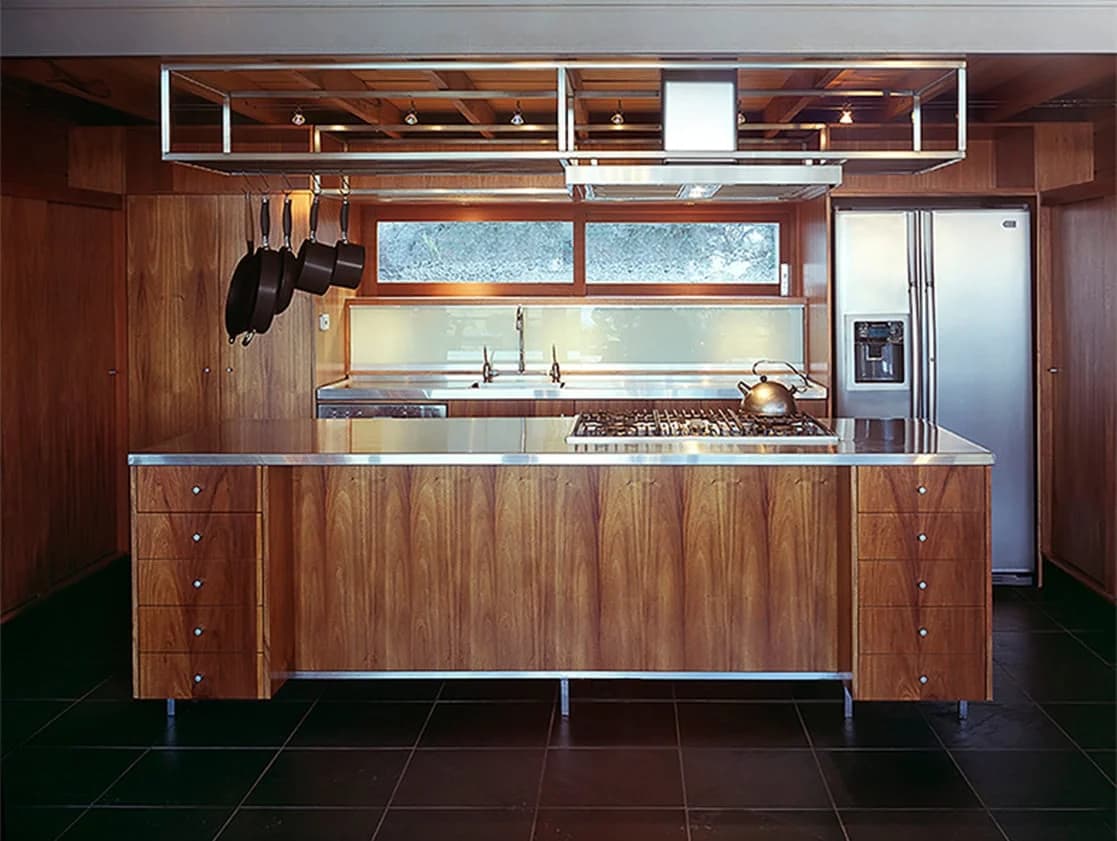Timber kitchen cabinets and island with metal worktops beneath an open overhead shelf.