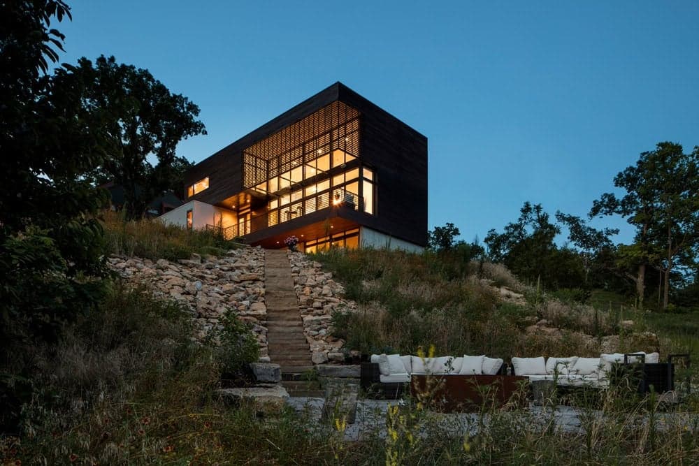 Illuminated timber-clad house above stone garden terraces and outdoor seating at dusk.