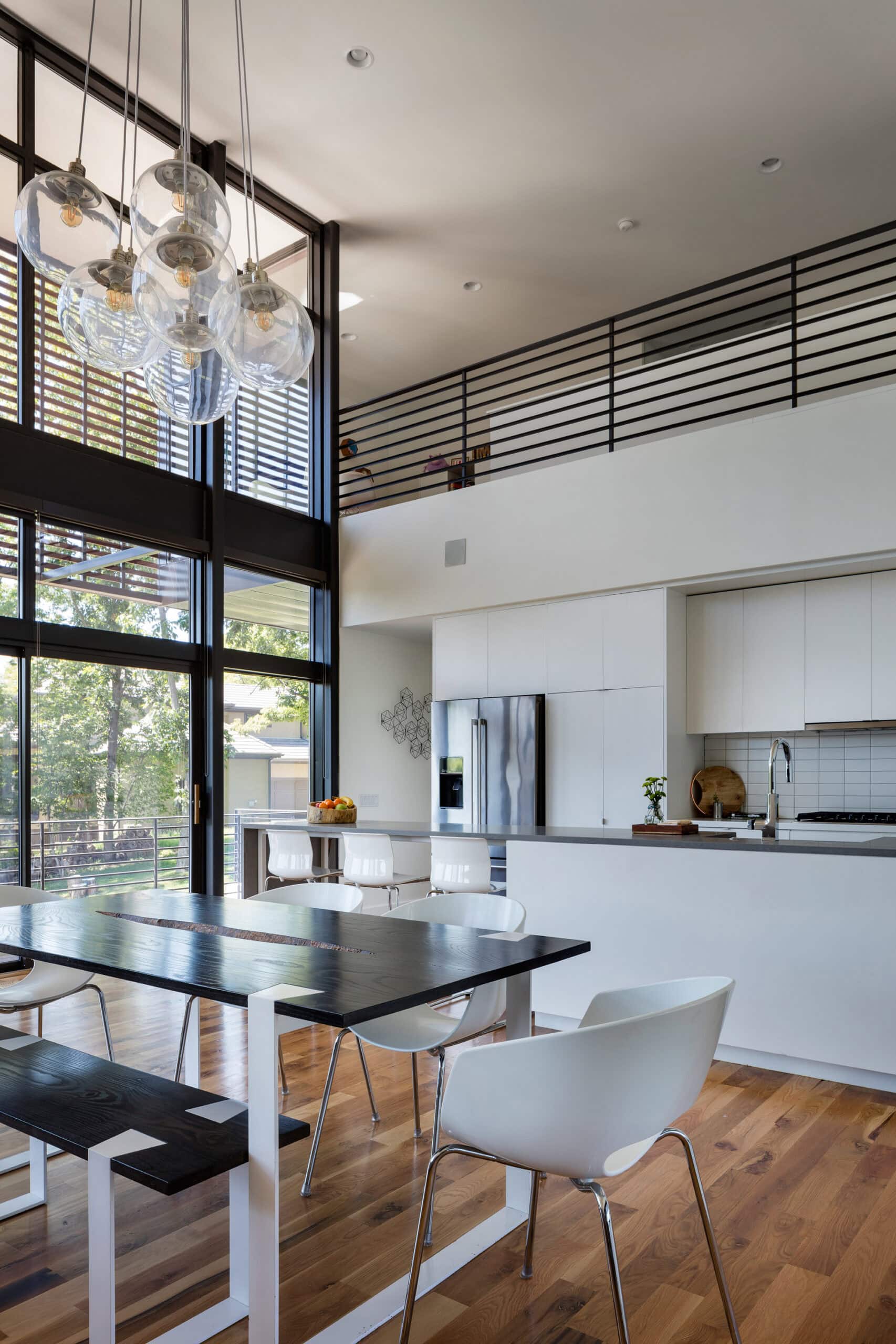White kitchen and dining table beneath pendant lights, double-height glazing and an upper-level balustrade.