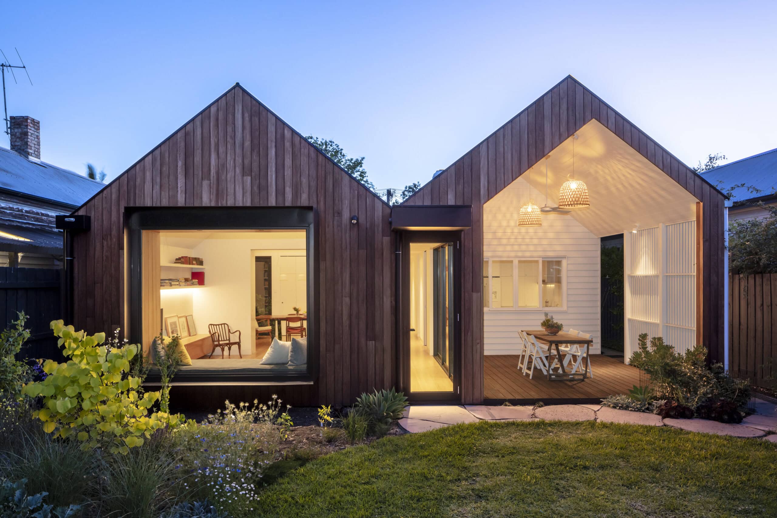Illuminated twin-gabled garden extension with a window seat and covered dining terrace at dusk.