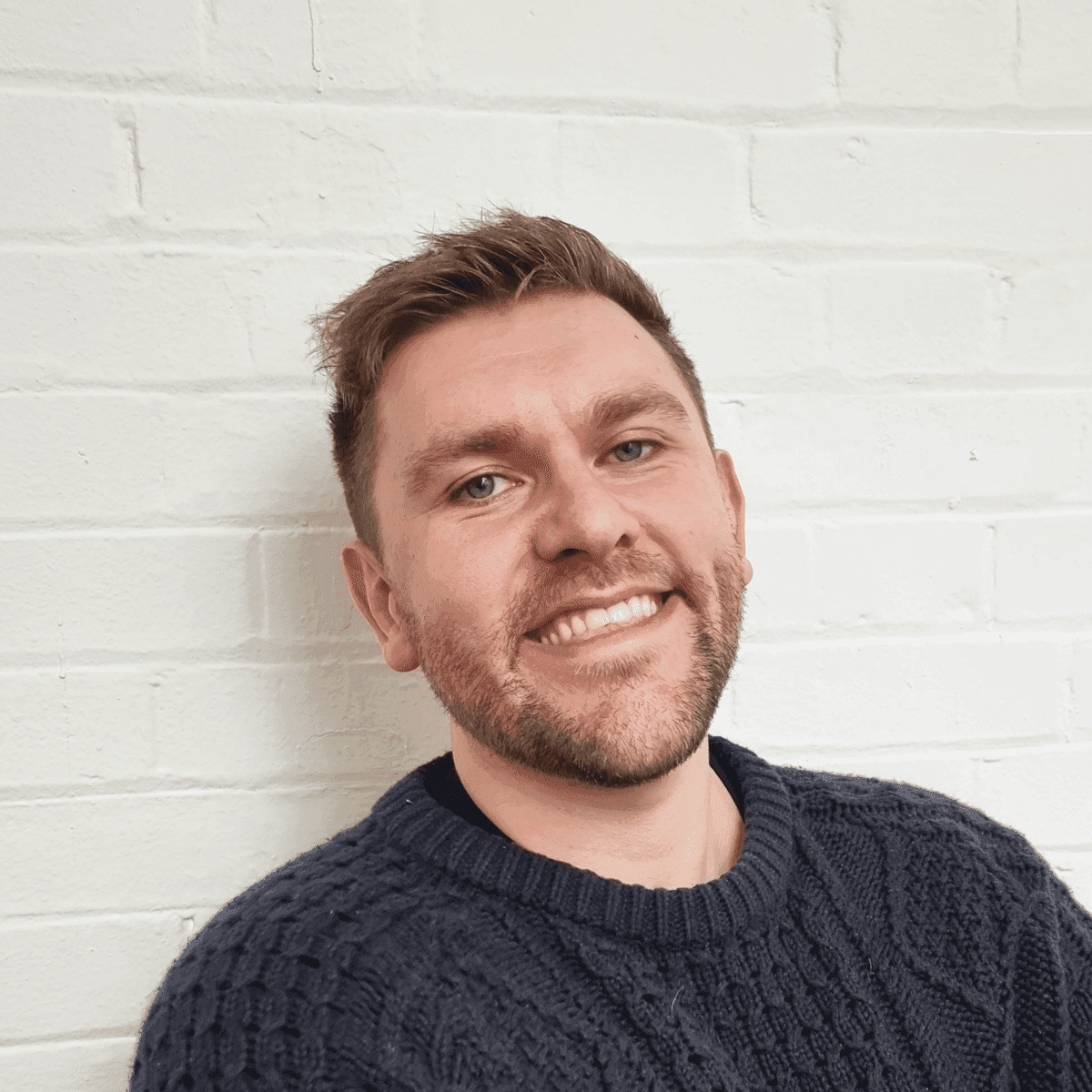 Stephen Drew smiling warmly in a cozy dark blue sweater against a white brick wall.