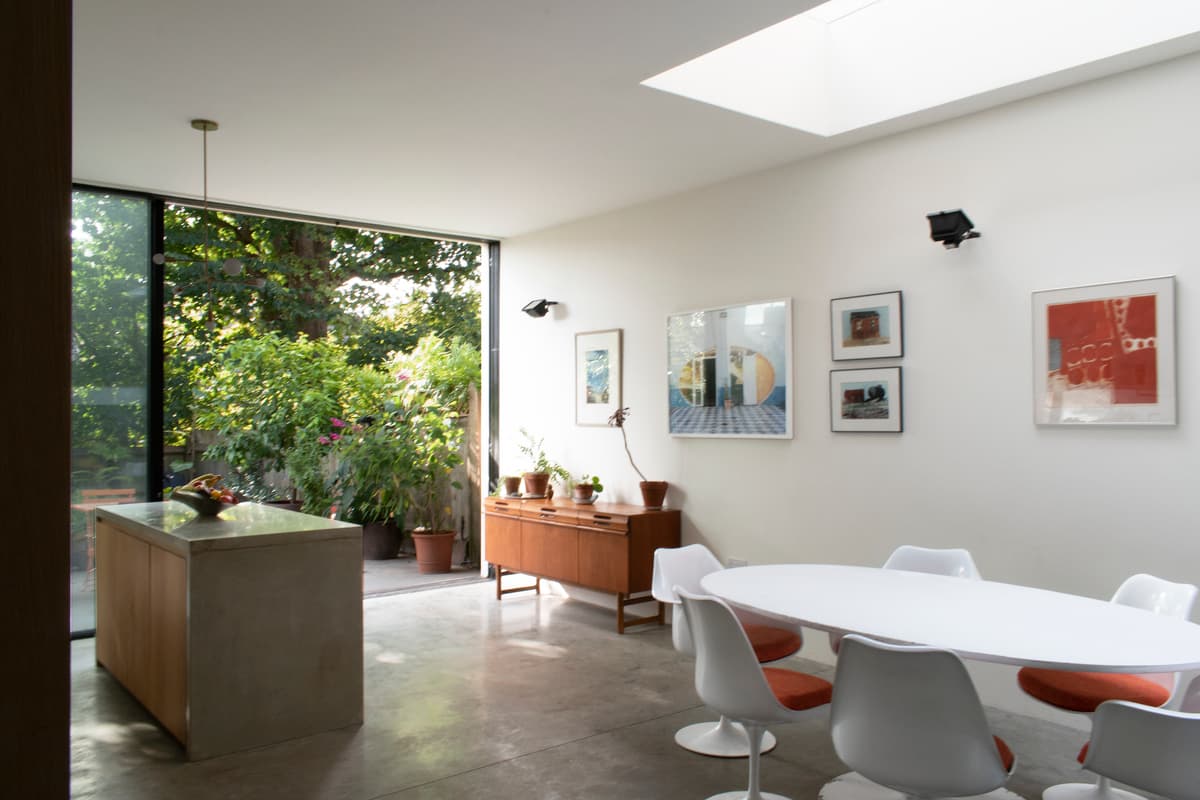 The dining table sits beside full-height garden glazing beneath a rooflight at Structuralist House.