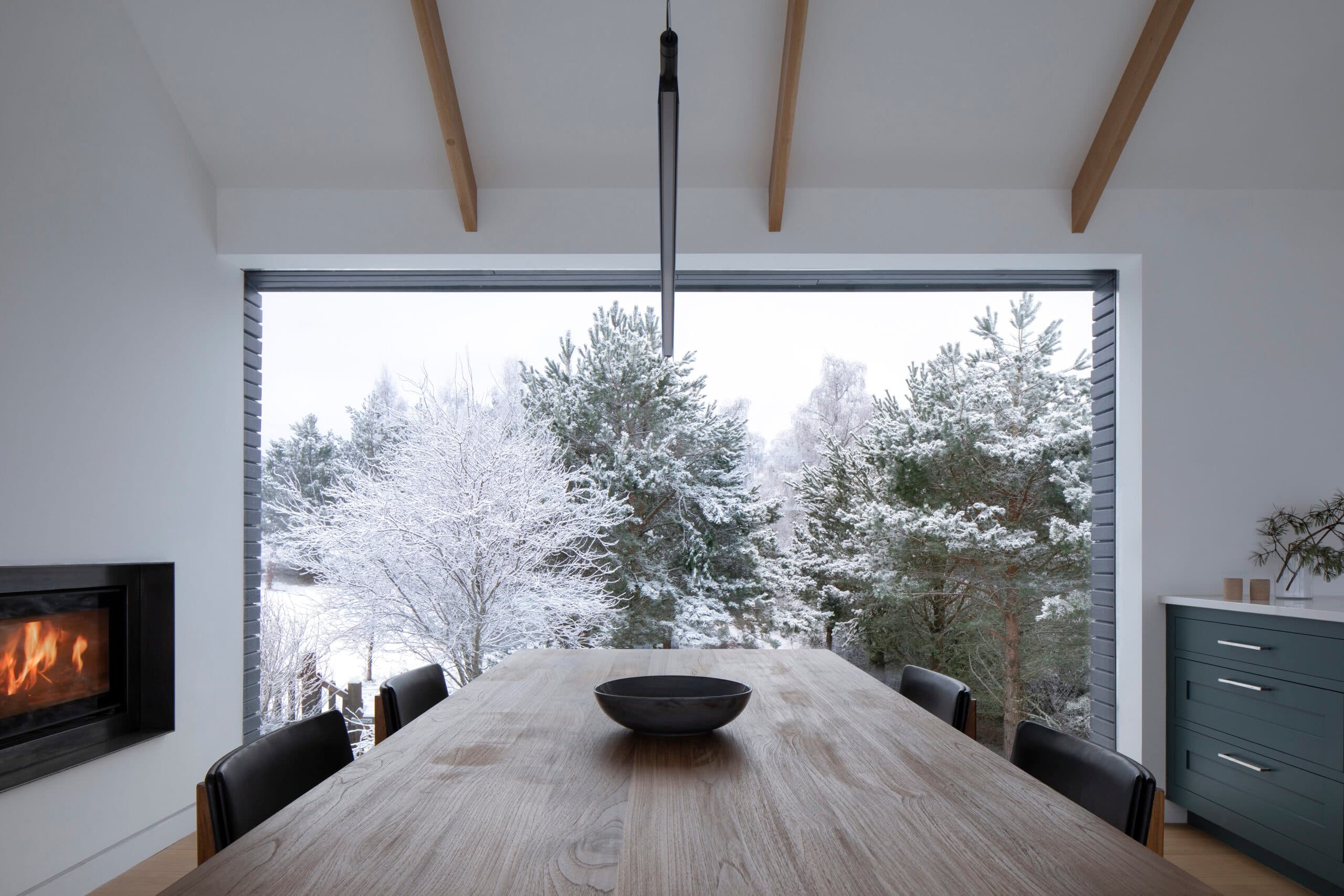 Dining table beneath exposed timber beams, facing a wide window onto snowy trees.