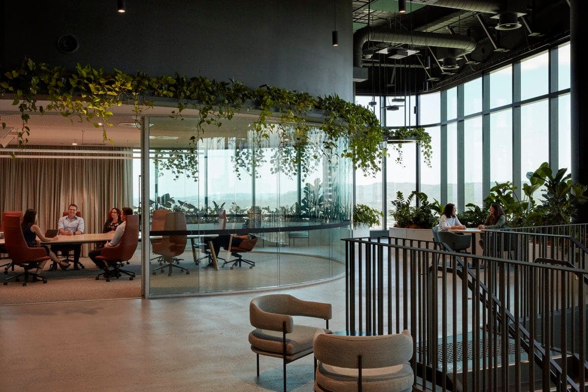 Modern open-plan office with glass-walled conference room, natural light, greenery, and stylish seating.