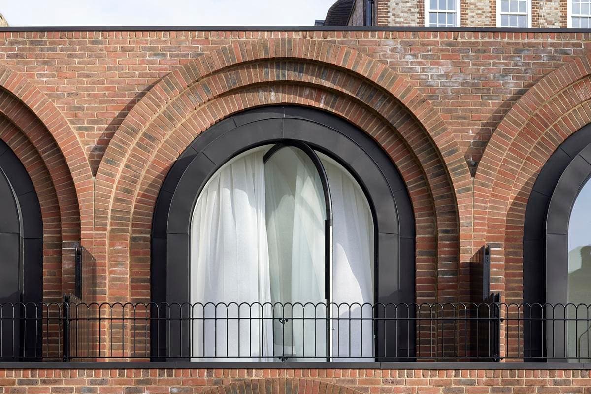 Arched window with dark frame, white curtains, and black railing on a red brick facade.