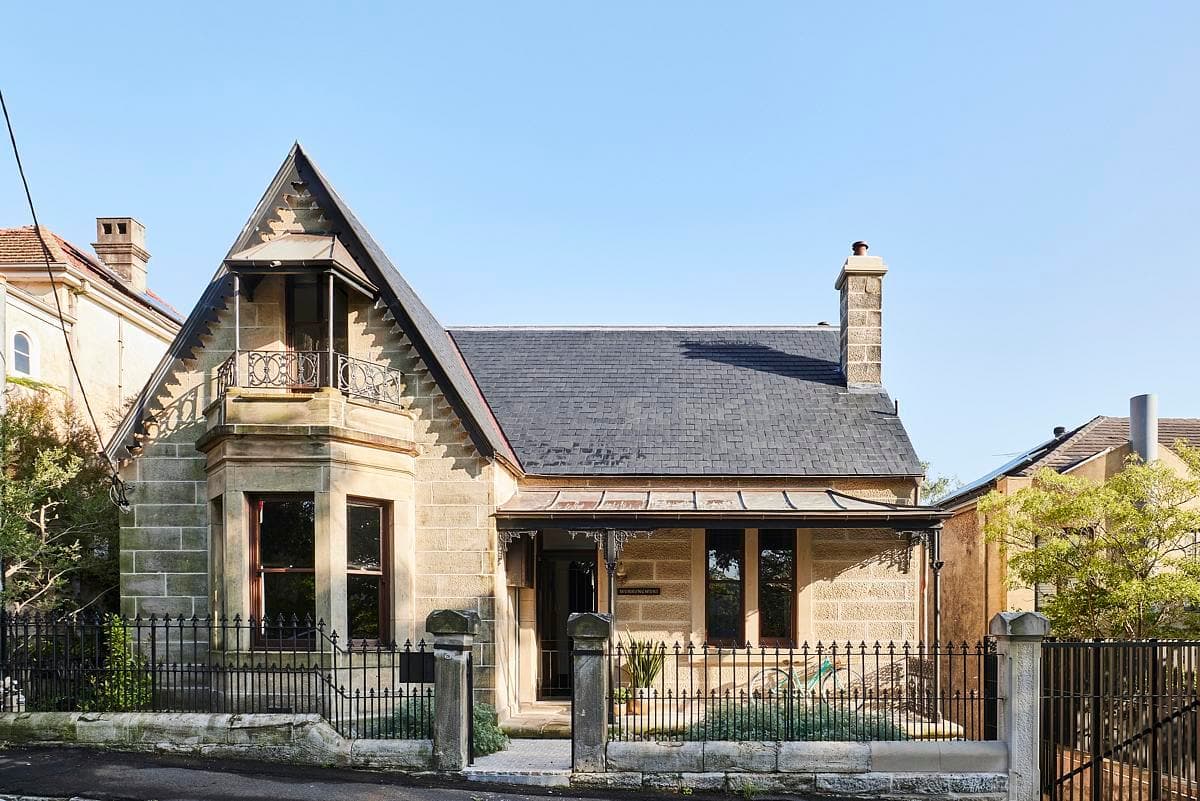 Victorian stone house with balcony, garden, and elegant architectural details against a clear blue sky.