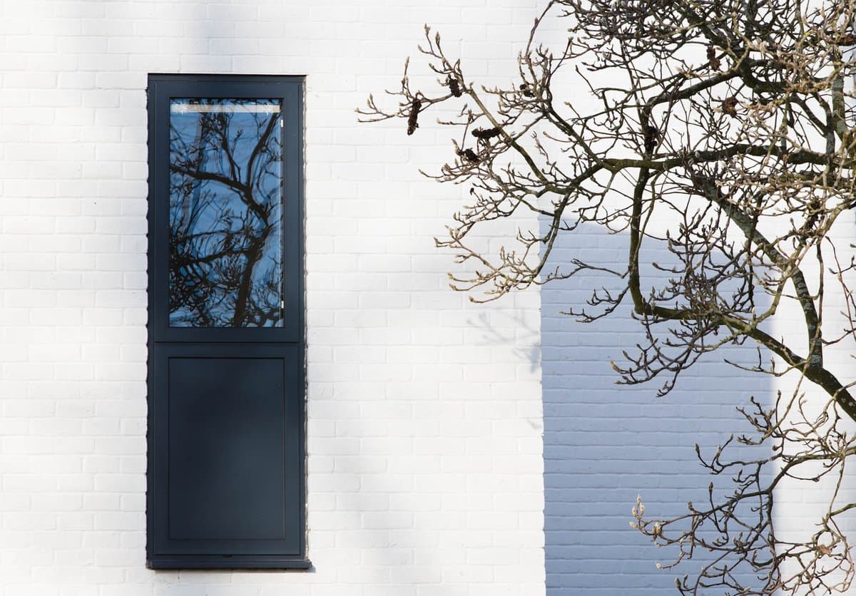 White brick wall with a tall, narrow window and reflective glass, adjacent to bare tree branches.