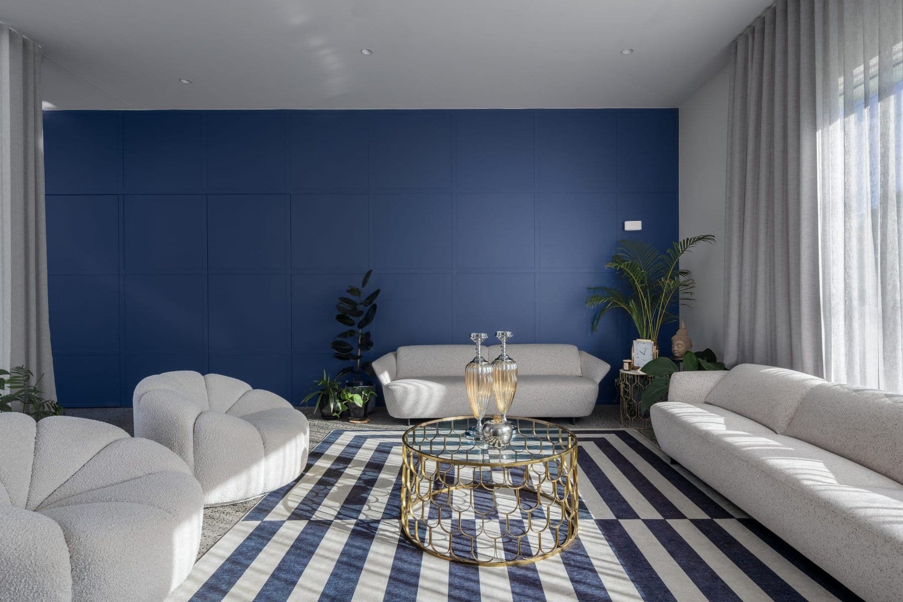 Modern living room with blue accent wall, white sofas, glass coffee table, and green plants.