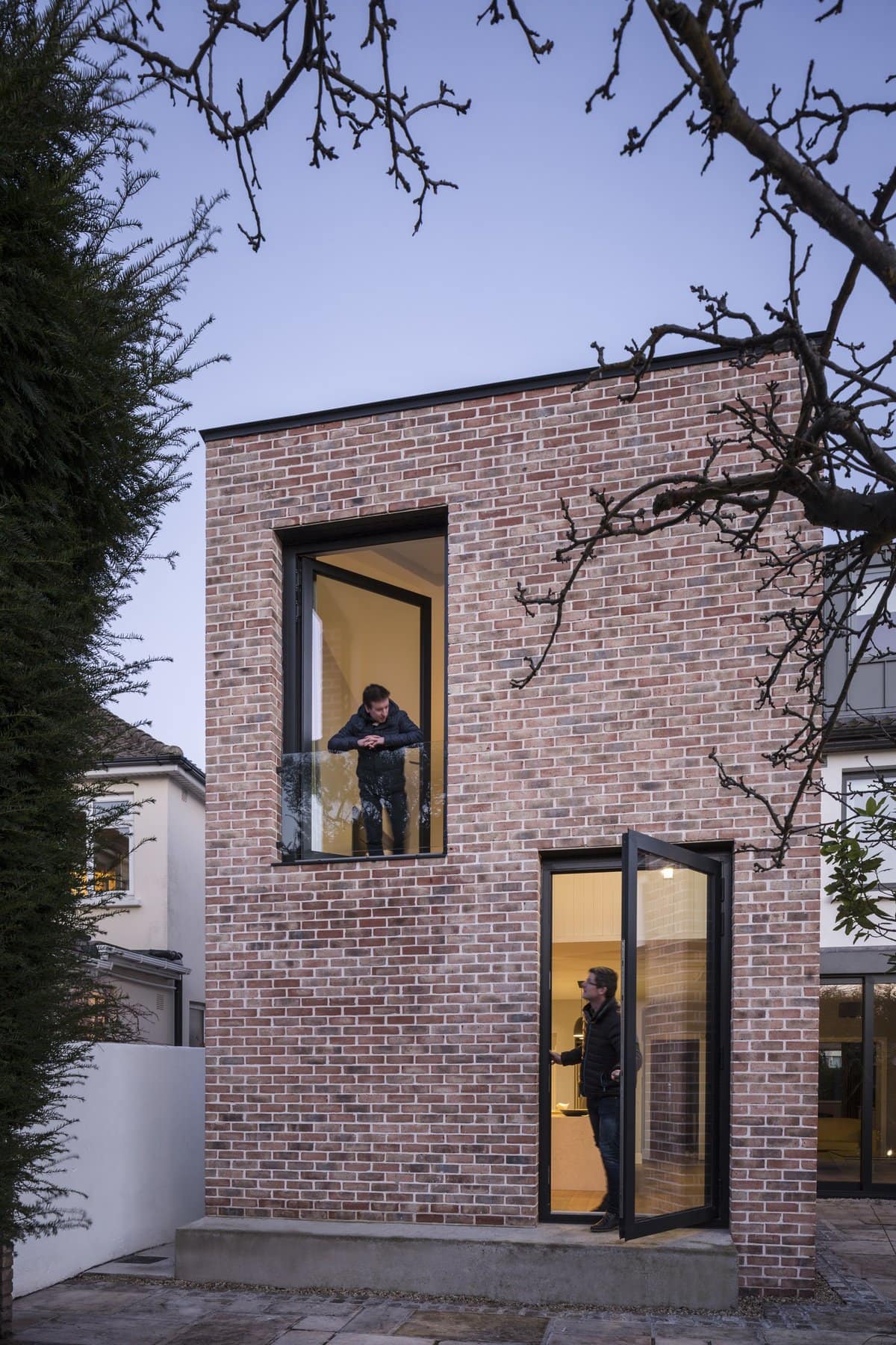 Modern two-story brick building with large windows, balcony, and individuals interacting indoors and outdoors.