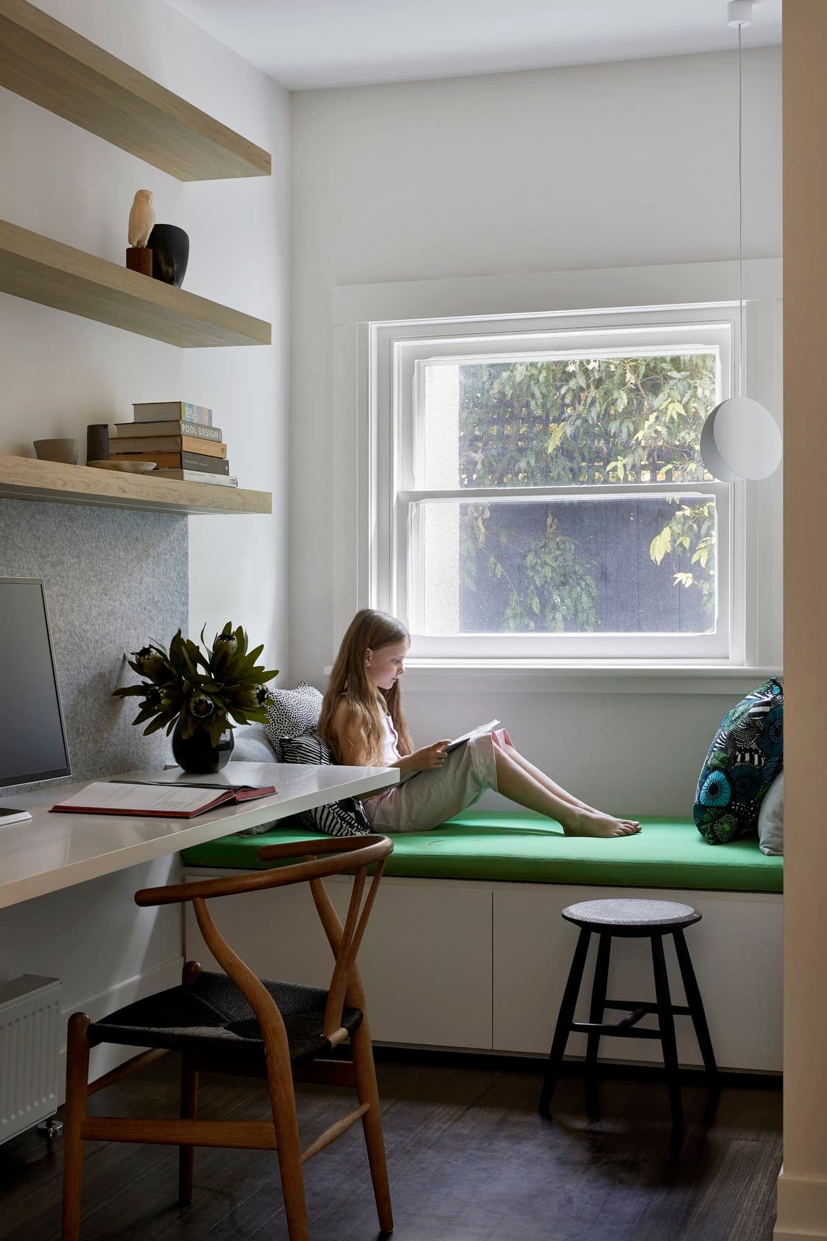 Cozy home office with natural light, green bench, minimalist desk, and serene greenery view.