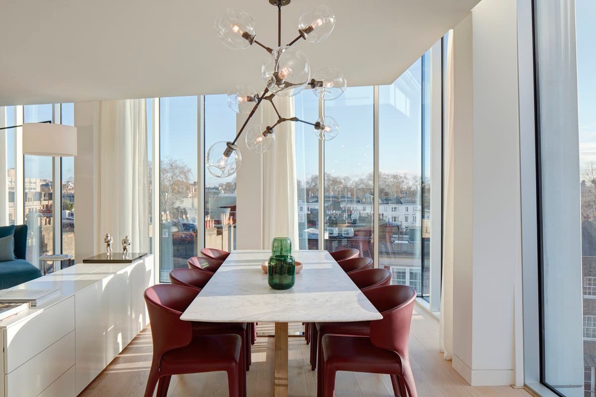 Modern dining area with marble table, red chairs, chandelier, and city view through large windows.