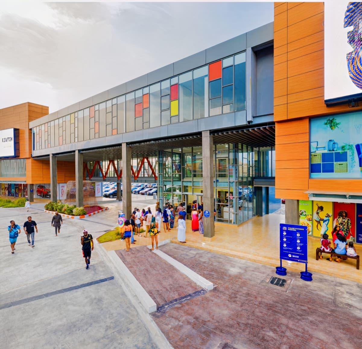 Modern commercial building with glass façade, orange accents, vibrant mural, landscaped plaza, and people.