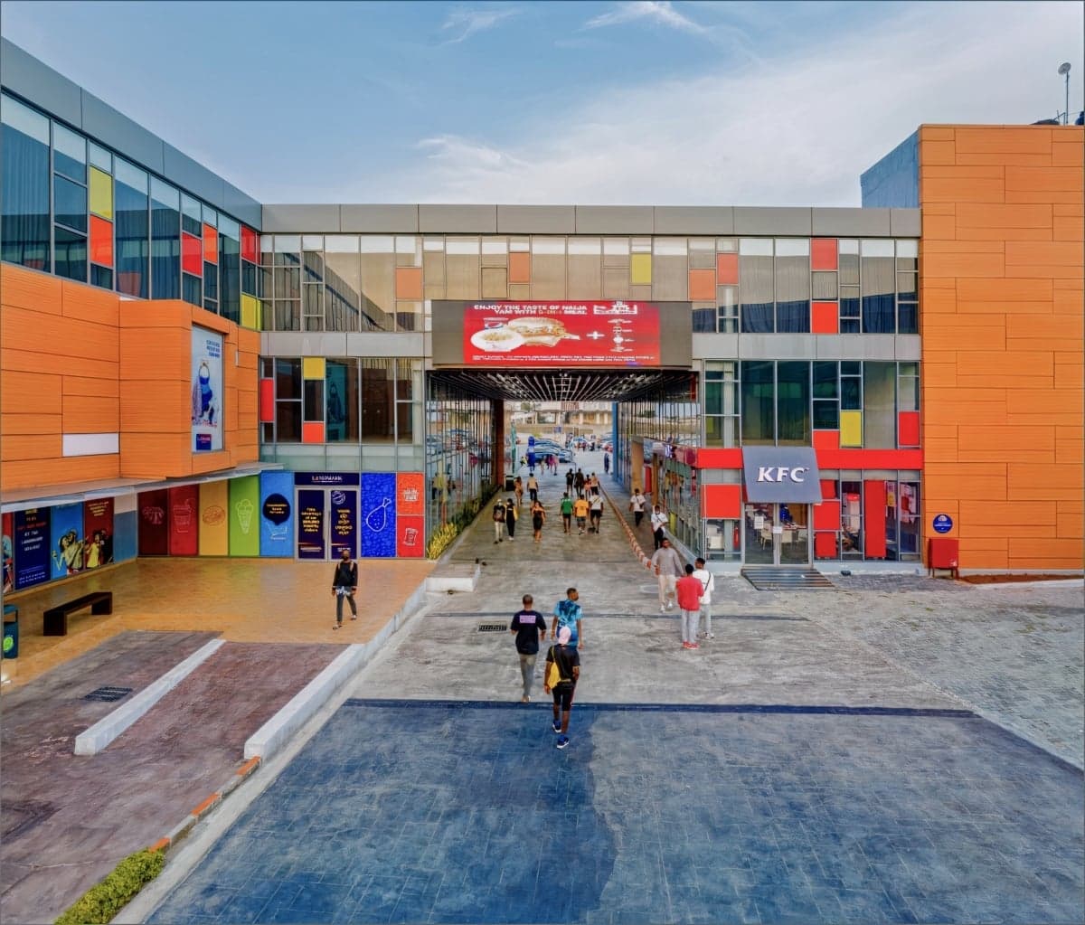 Modern shopping mall entrance with colourful facade, paved area, and prominent KFC outlet.