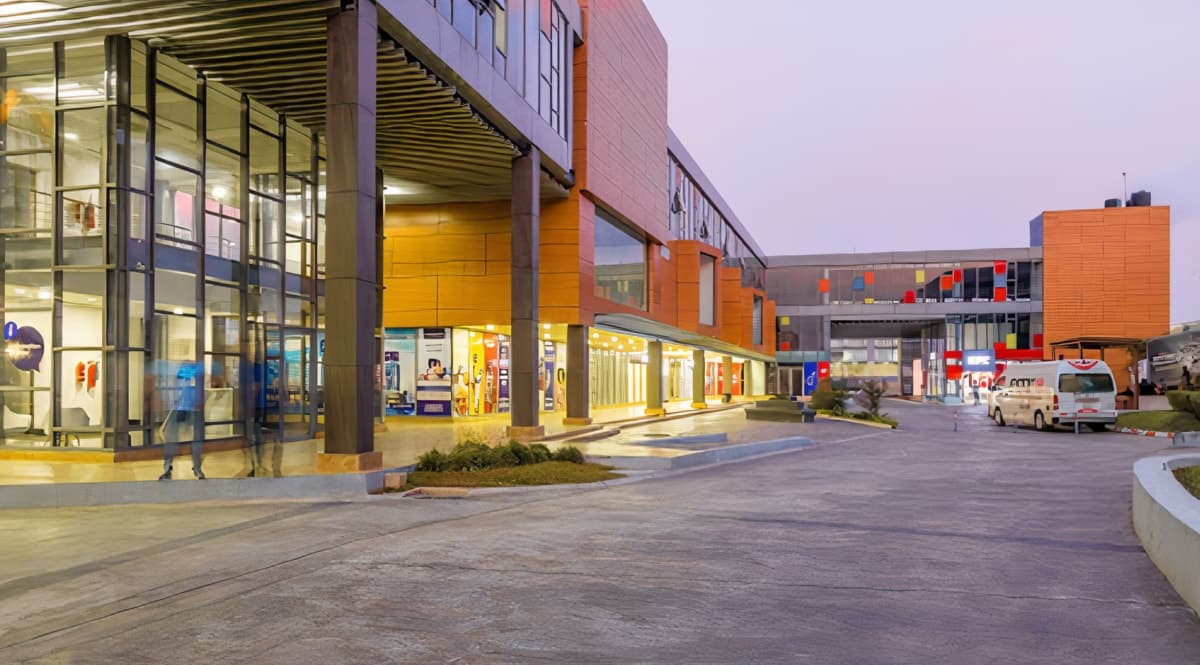 Modern commercial building at dusk with glass facade, shops, overhanging roof, and landscaped driveway.