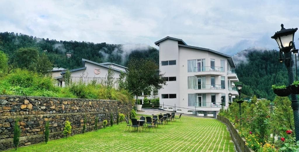 Modern white hotel amidst lush greenery, with outdoor seating, misty backdrop, and elegant streetlamp.