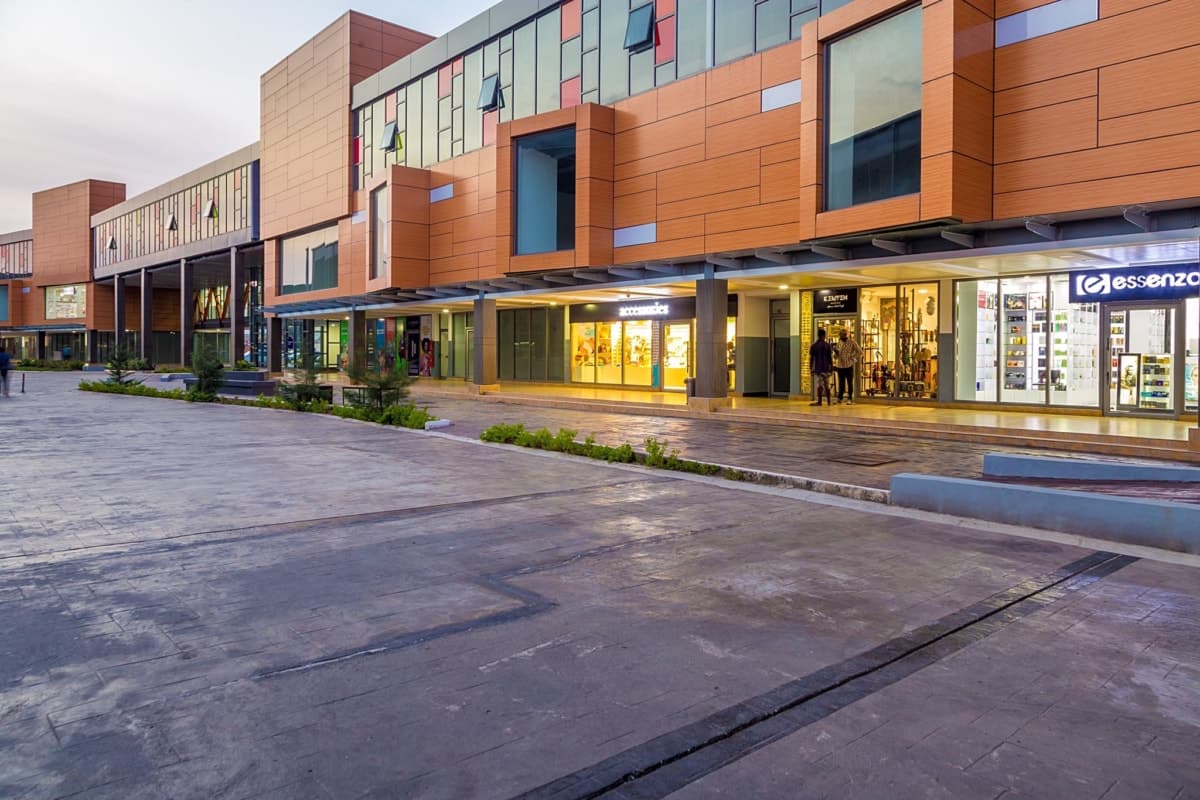 Modern shopping area with paved path, green shrubs, and contemporary glass-fronted stores.