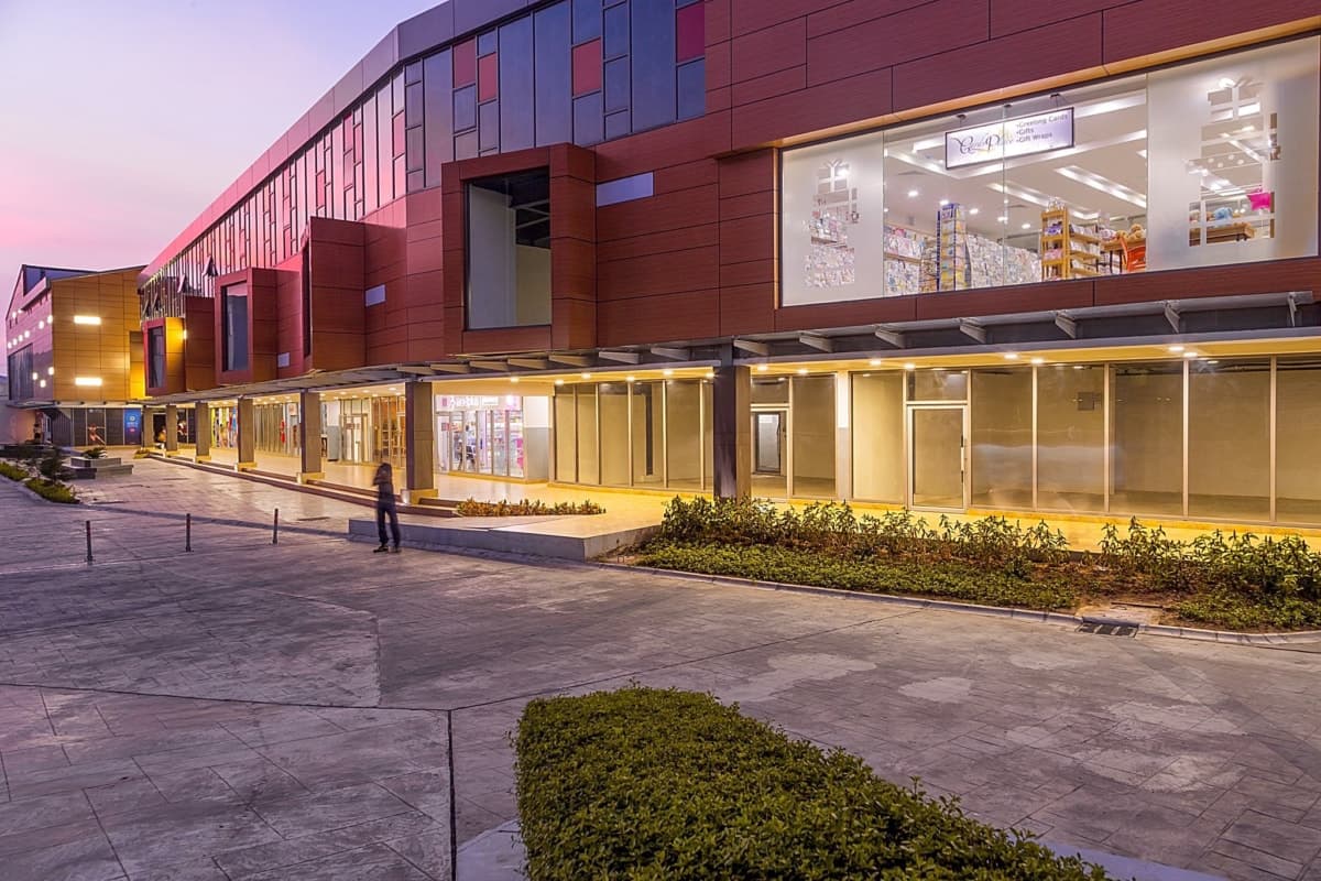 Modern shopping mall exterior at dusk with illuminated storefronts and pedestrian walkway.