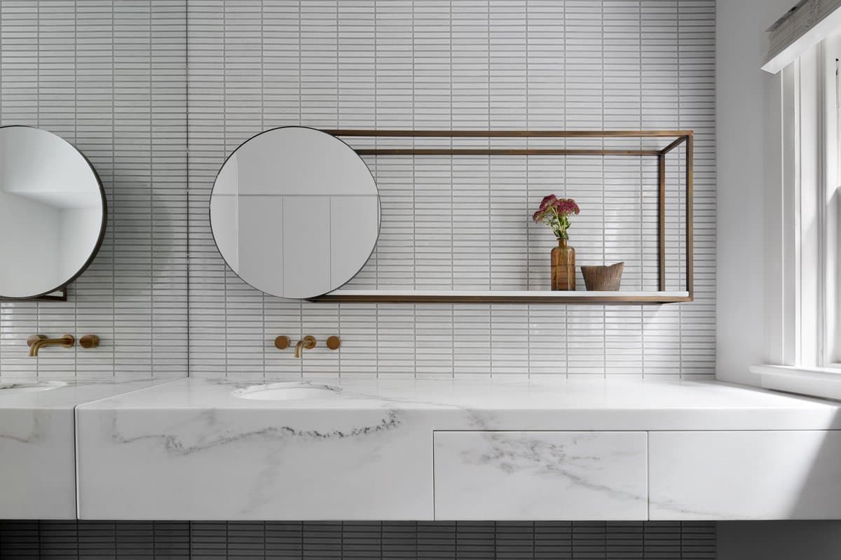 Minimalist bathroom with white tiles, brass fixtures, twin sinks, and natural light.