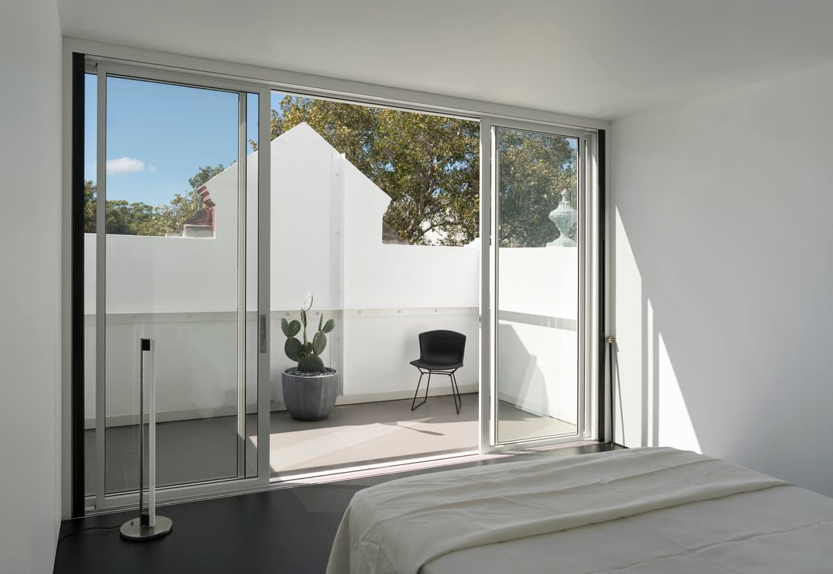 Modern minimalist bedroom with glass door, natural light, white walls, dark flooring, and outdoor balcony.
