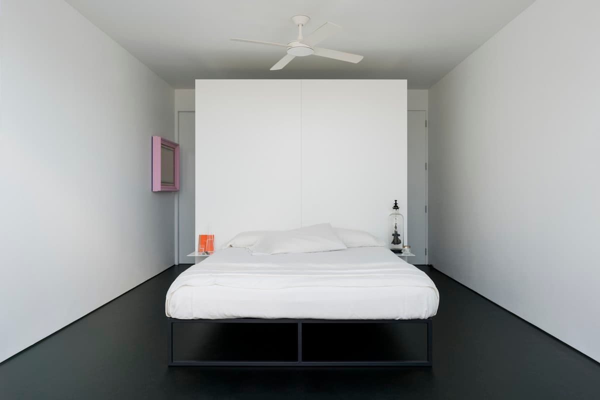 Minimalist bedroom with black bed frame, white bedding, pink art, and black nightstand with lamp.