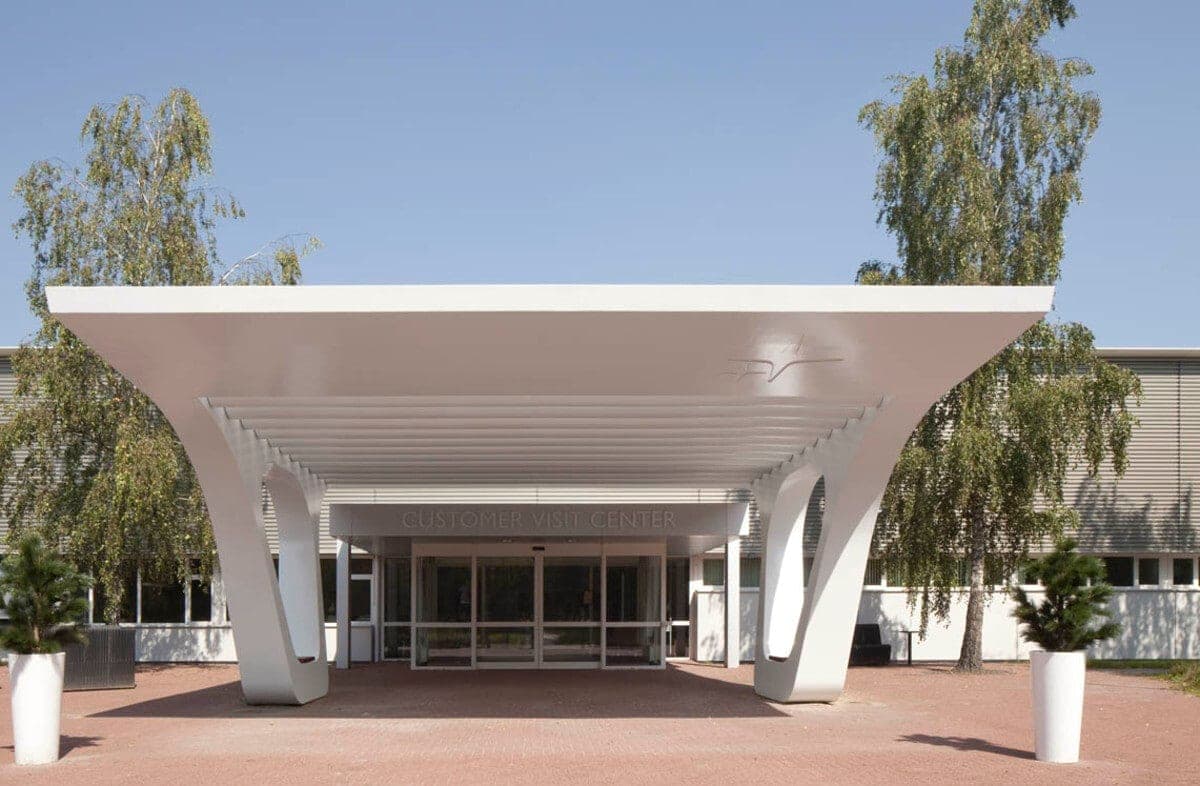 Modern entrance canopy with white columns, glass doors, red brick paving, and green plants.