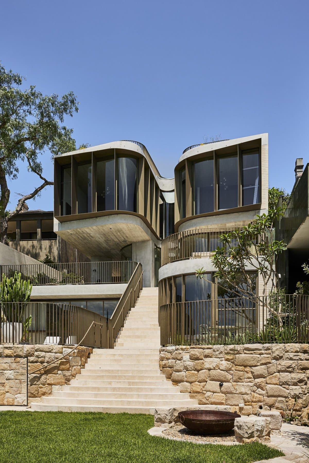 Curved house facade with glazing, terraces and surrounding planting.